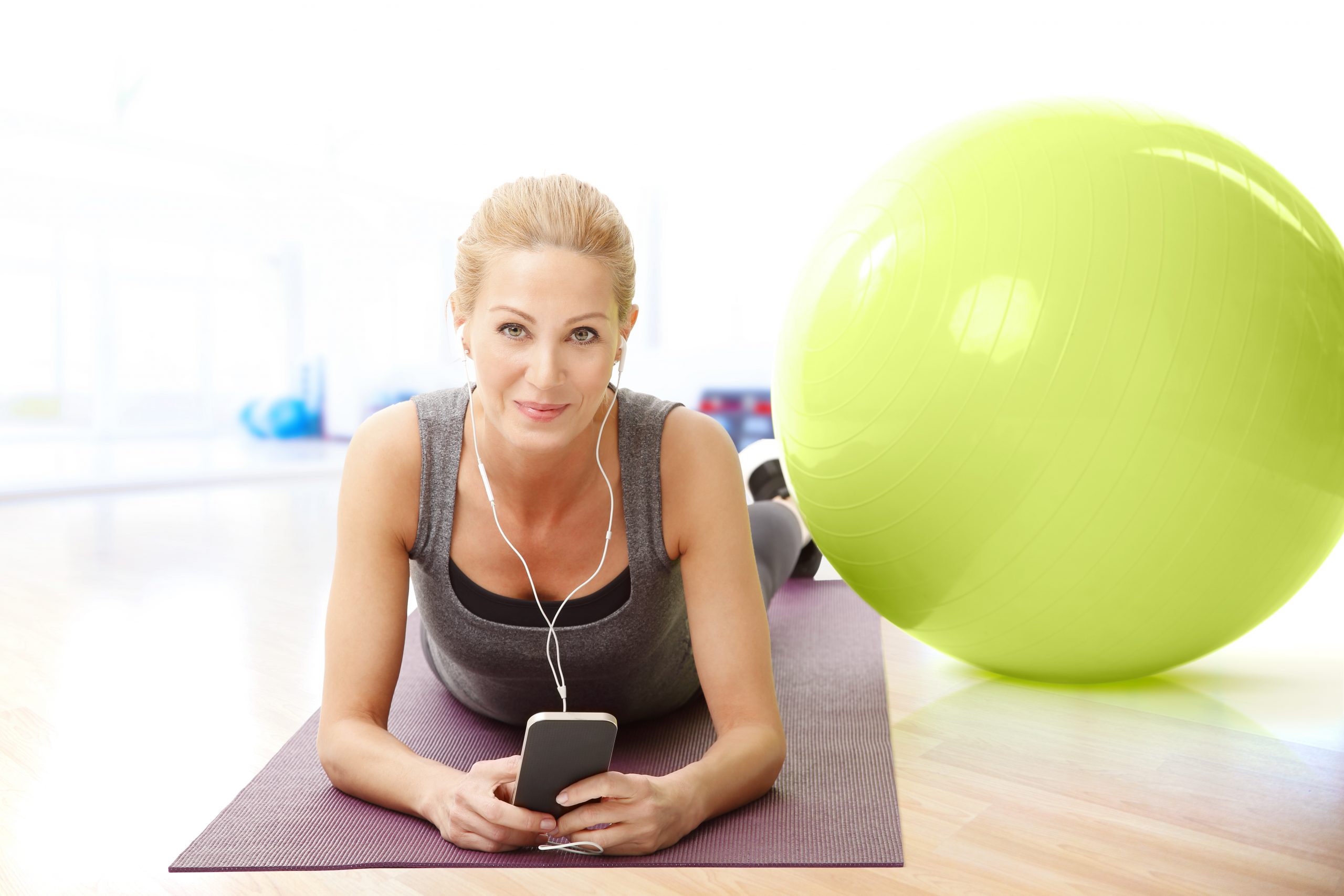 Portrait of smiling middle age woman lying on exercise mat and listening music with mobile phone while relaxing after fitness workout.